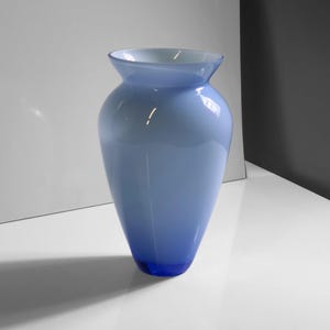 Könnte beinhalten: Eine hohe, elegante blaue Glasvase mit ausgestelltem Rand. Die Vase hat eine Farbverlaufsfarbe, die von einem hellen Blau oben zu einem tiefen Blau am Boden übergeht. Die Vase ist leer und steht auf einer weißen Oberfläche.