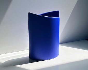 Może przedstawiać: Kobaltowy, cylindryczny wazon z unikalną, złożoną górą. Wazon wykonany jest z matowego materiału i stoi na białej powierzchni. Oświetlenie tworzy mocny cień, podkreślający kształt i fakturę wazonu.