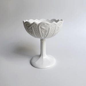 Puede incluir: Un cuenco blanco de vidrio de leche con pedestal, borde festoneado y diseño en relieve intrincado. El cuenco está sobre un fondo blanco.