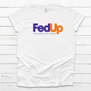 May include: White t-shirt with the word "FedUp" in purple and orange, mimicking a well-known logo, with the text "with everything and everyone" below. The shirt is displayed on a white wooden surface.