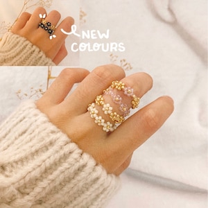 May include: A hand wearing three beaded rings. The rings are made of small white beads with gold accents and a pink beaded ring. The text "NEW COLOURS" is written on the image.