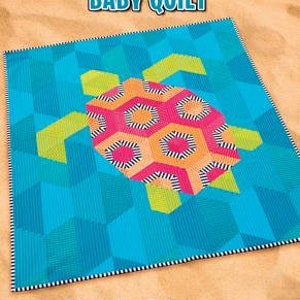 May include: A baby quilt featuring a colorful geometric turtle design on a blue background. The quilt is bordered with a white and black stripe. The text "TULLY THE TURTLE BABY QUILT" is at the top of the quilt. The text "Krista Moser The Quilted Life" is at the bottom of the quilt. The text "QUILT SIZE 40 1/2" x 47" is at the bottom of the quilt.
