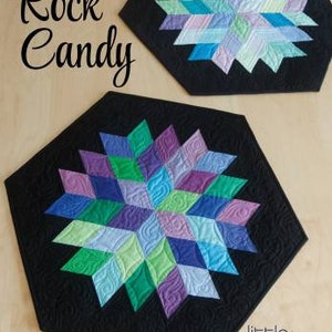 May include: A black hexagon-shaped quilted table runner with a colorful geometric design in shades of purple, green, blue, and pink. The design is called "Rock Candy" and is by Little Jaybird Quilts.