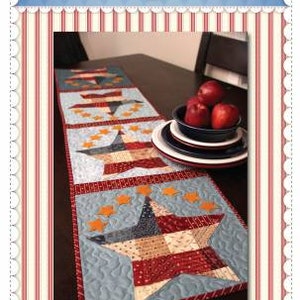 May include: A patchwork table runner with a patriotic theme. The runner features a blue background with red, white, and blue stars and stripes. The text "Patchwork Patriotic Table Runner" is printed below the runner.