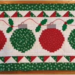 May include: A green and red Christmas table runner with a white background and a string of ornaments with holly leaves.