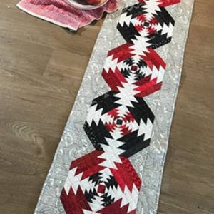 May include: A long table runner with a white background and a repeating pattern of red, black, and white geometric shapes. The runner is made of fabric and has a quilted texture.