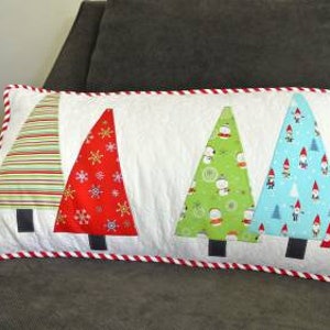 May include: A white pillow with a red and white candy cane trim features five colorful Christmas trees. The trees are made of fabric and have different patterns, including stripes, snowflakes, snowmen, gnomes, and polka dots.