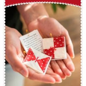 May include: Two small red and white quilted squares with a pocket for a prayer card. The squares are held in a person's hands. The text on the card reads "The Pocket Prayer Quilt".