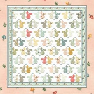 May include: A quilt with a repeating pattern of bunny shapes in various colors, including pink, blue, and yellow. The quilt is framed with a green border and set against a peach background with small mushroom and leaf designs. The text "Bunny Town" and "by Coach House Designs" are displayed.