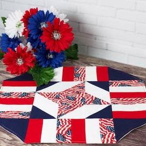 May include: A patriotic table runner with a star design, featuring red, white, and blue colors and American flag patterns. The runner is placed on a wooden surface, with a bouquet of red, white, and blue flowers in the background.
