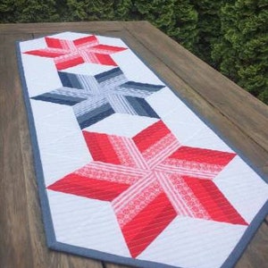 May include: A white table runner with a gray border features three red, white, and blue star designs. The stars are layered and quilted, creating a dimensional effect.
