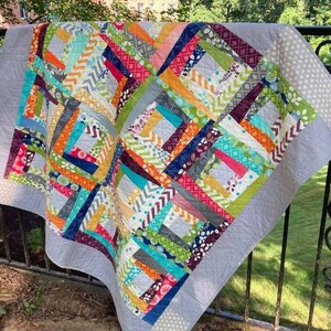 May include: A colorful patchwork quilt with a variety of fabrics in shades of blue, green, orange, red, and gray. The quilt is made up of squares and rectangles, and the fabric is sewn together in a random pattern. The quilt is hanging on a fence.