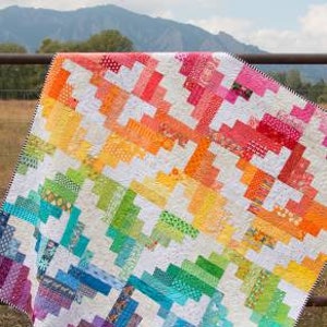 May include: A colorful quilt pattern called "Mapleton Avenue" by Sassafras Lane Designs. The quilt is made up of squares in a rainbow of colors, with white squares interspersed throughout. The quilt is hanging on a fence in front of a mountain.