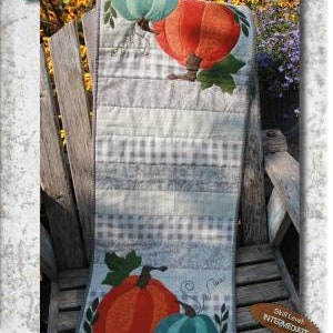 May include: A quilt-as-you-go table runner featuring a pumpkin patch design. The runner is made of gray and white fabric with a checkered pattern. The pumpkins are orange, green, and teal. The text "Pumpkin Patch" and "Finish in One Day!" is printed on the runner. The text "P251" and "Intermediate Level" is also printed on the runner.