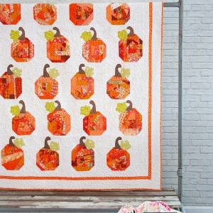 May include: A white quilt with a patchwork design featuring orange and brown pumpkins. The quilt is hanging on a white brick wall with a metal rod. The text "Patch Work" and "Fresh designs from a Wyoming farm." is printed below the quilt.