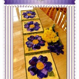 May include: A patchwork table runner with a pansy flower design in shades of purple and yellow. The runner is made of fabric squares sewn together to create a quilted effect. The text "Patchwork Pansy Table Runner" is printed at the bottom of the image.