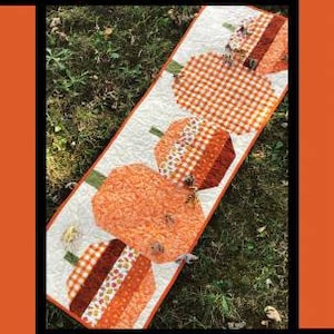 May include: A quilted table runner with a pumpkin theme. The runner features five pumpkins in shades of orange and brown with a white background. The runner is titled "Pumpkin Hollow" and is by Tricia Lynn Maloney for Villa Rosa Designs.