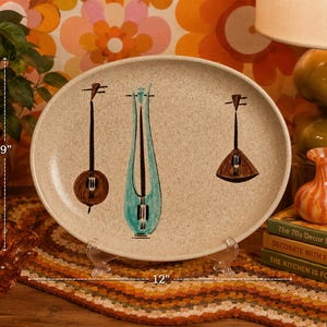 May include: An oval serving platter with a speckled beige surface, featuring three stylized musical instrument illustrations in brown and teal. The platter is 12 inches long and 9 inches wide. The platter is displayed on a clear stand.