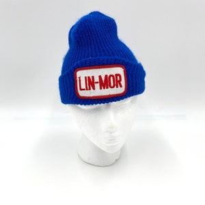May include: A blue knitted beanie with a red and white patch that says "LIN-MOR" on it.