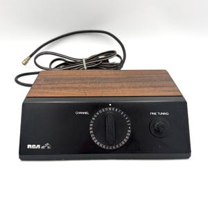 May include: A vintage RCA black and brown TV tuner with a channel dial and fine tuning knob. The top is wood-grained, and a cable extends from the back. The RCA logo is visible on the front.