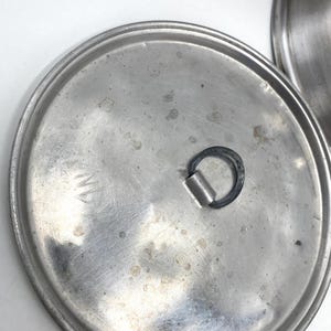 Op de afbeelding: Close-up van een roestvrijstalen pannendeksel met een handvat. Het deksel is cirkelvormig met een licht getextureerd oppervlak en een donker handvat. Het deksel heeft wat slijtage, met zichtbare vlekken en markeringen.
