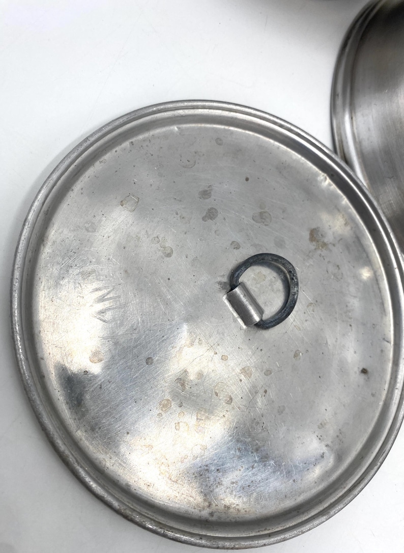 Op de afbeelding: Close-up van een roestvrijstalen pannendeksel met een handvat. Het deksel is cirkelvormig met een licht getextureerd oppervlak en een donker handvat. Het deksel heeft wat slijtage, met zichtbare vlekken en markeringen.