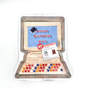 May include: A silver computer-shaped cake pan with a blue screen that says "Happy Birthday Mac". The pan has a keyboard with red, orange, blue, and white keys.