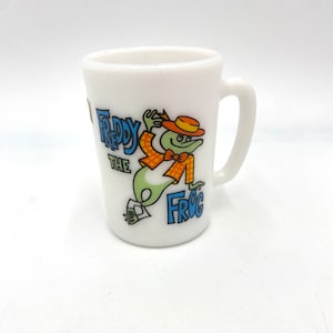 May include: White ceramic mug with a cartoon frog wearing a hat and a bow tie. The frog is green with orange and red accents. The text "Freddy the Frog" is printed on the mug.