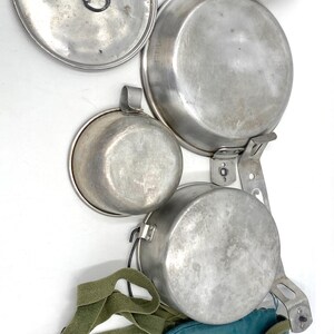 Op de afbeelding: Een vintage kampeerkookset met verschillende aluminium potten, pannen en een deksel. De set bevat een kleine beker, een groene canvas draagtas en een groene riem. De items zijn gerangschikt op een witte achtergrond.