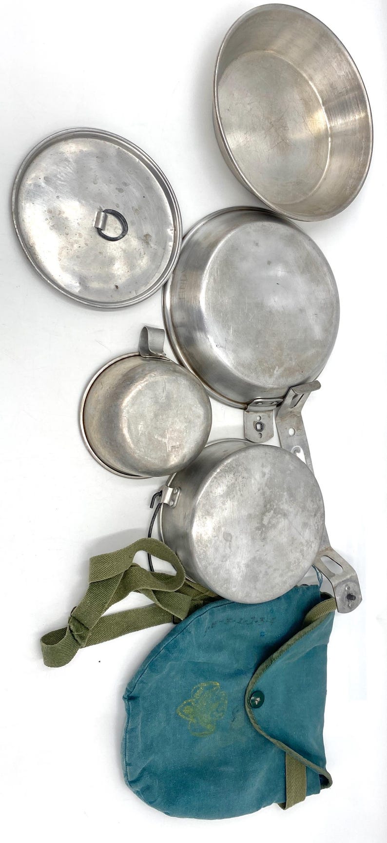 Op de afbeelding: Een vintage kampeerkookset met verschillende aluminium potten, pannen en een deksel. De set bevat een kleine beker, een groene canvas draagtas en een groene riem. De items zijn gerangschikt op een witte achtergrond.