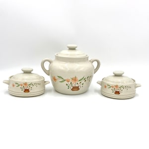 Op de afbeelding: Een set van drie crèmekleurige keramische serveerschalen met deksels. Elk stuk heeft een bloemenontwerp met perzikroze bloemen en groene bladeren. De grootste pot heeft twee handvatten. Ideaal voor het serveren van soepen, stoofschotels of andere gerechten.