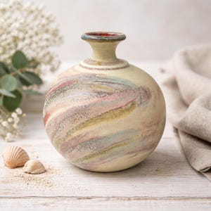 May include: A round ceramic vase with a small neck and a wide body. The vase has a cream-colored base with swirling patterns of pink, green, and yellow. The rim is red with a blue accent. Two seashells and a white flower arrangement are in the background.