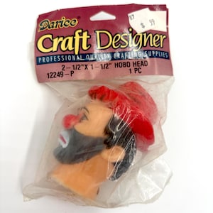 May include: A Craft Designer Hobo Head craft supply in a clear plastic bag. The hobo head is approximately 6.35cm x 3.81cm and features a red hat, black beard, and a red nose. The packaging includes the Darice logo and product information.