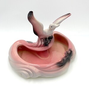 以下が含まれることがあります： 縁にカモメの置物が置かれたピンクの陶器製の皿。皿は波のようなデザインで、薄いピンク色に黒のアクセントが施されています。