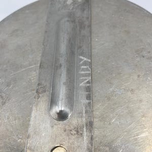 Op de afbeelding: Close-up van een metalen deksel met een handvat. Het deksel is cirkelvormig met een geborstelde metalen afwerking. Een rechthoekig handvat is aan het deksel geklonken, met het woord "NUX" erin ge&euml;tst. Een klein embleem is zichtbaar op het handvat.