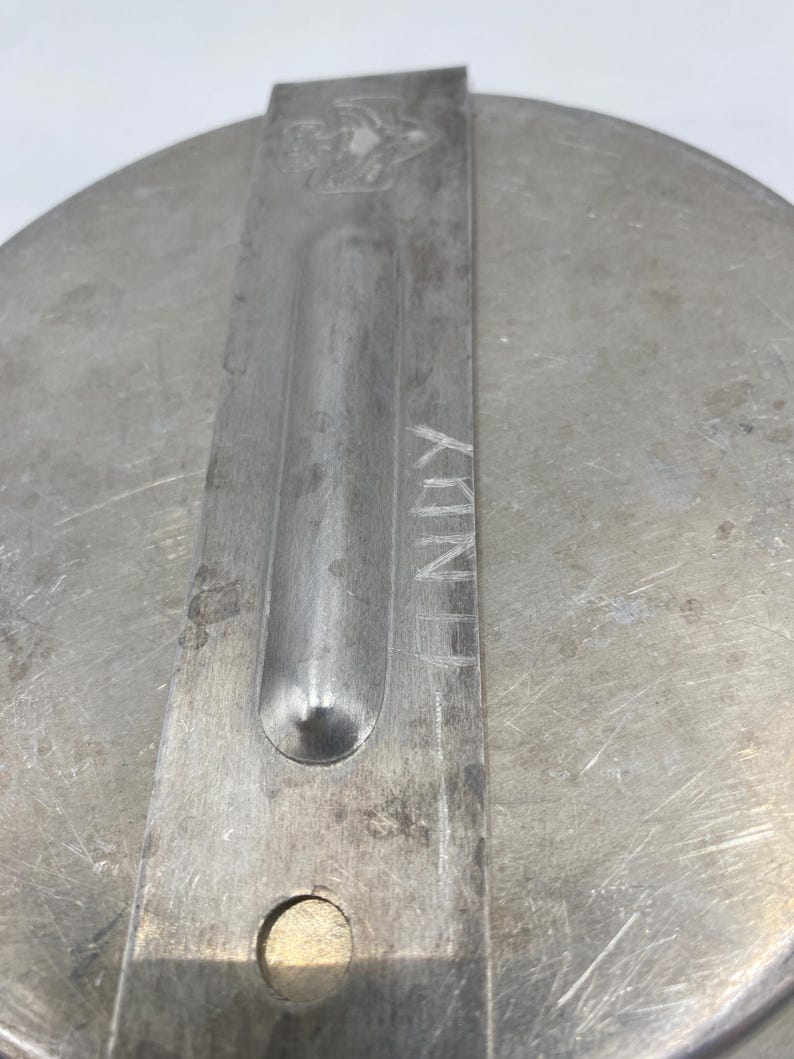 Op de afbeelding: Close-up van een metalen deksel met een handvat. Het deksel is cirkelvormig met een geborstelde metalen afwerking. Een rechthoekig handvat is aan het deksel geklonken, met het woord "NUX" erin ge&euml;tst. Een klein embleem is zichtbaar op het handvat.