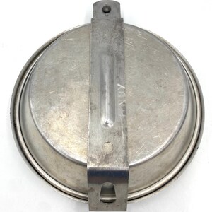 Op de afbeelding: Een vintage, ronde, zilverkleurige messkit met een scharnierend handvat en een sluiting. De kit lijkt van metaal te zijn en vertoont tekenen van slijtage. Het woord "HENRY" is op het handvat gestempeld.