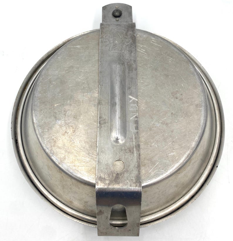 Op de afbeelding: Een vintage, ronde, zilverkleurige messkit met een scharnierend handvat en een sluiting. De kit lijkt van metaal te zijn en vertoont tekenen van slijtage. Het woord "HENRY" is op het handvat gestempeld.