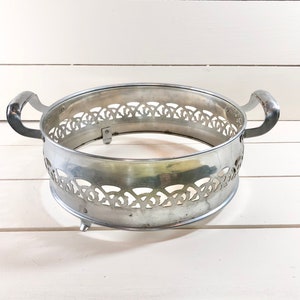 Op de afbeelding: Een zilverkleurige metalen serveerschaalstandaard met twee gebogen handvatten. De standaard heeft een decoratief opengewerkt ontwerp aan de zijkanten en drie kleine voetjes. Ontworpen om een ronde schaal te bevatten.