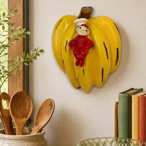 May include: A yellow ceramic wall plaque shaped like a bunch of bananas with a brown stem. A small, red-clad figure with a white chef hat is attached to the center of the bananas. The piece features dark brown vertical accents along the fruit.