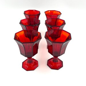 May include: Six vintage red glass goblets with a faceted design. The goblets have a hexagonal base and a flared, octagonal bowl. The stem and base are also hexagonal. The glasses are arranged in two rows of three on a white background.