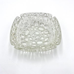 Peut inclure: Un cendrier en verre transparent avec un design texturé en forme de bulles. Le cendrier est carré avec des coins arrondis.