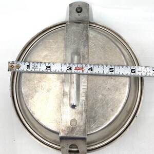 Op de afbeelding: Een vintage, ronde, zilverkleurige metalen messkit met een scharnierend handvat en een sluiting. De kit meet ongeveer 15 cm in diameter. Het handvat heeft een reli&euml;f ontwerp. Een meetlint ligt erop.