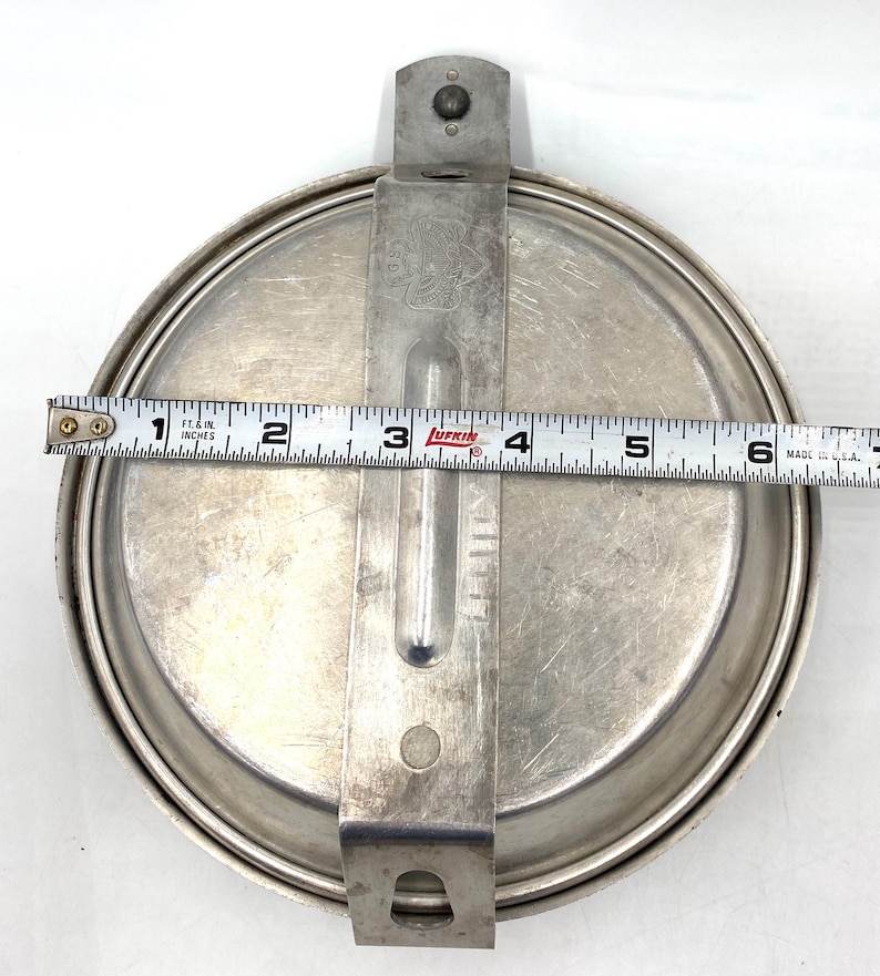 Op de afbeelding: Een vintage, ronde, zilverkleurige metalen messkit met een scharnierend handvat en een sluiting. De kit meet ongeveer 15 cm in diameter. Het handvat heeft een reli&euml;f ontwerp. Een meetlint ligt erop.