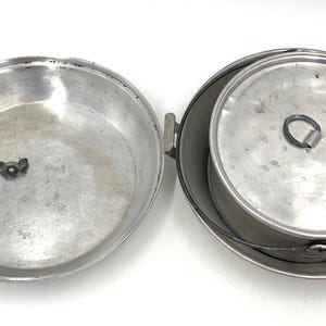 Op de afbeelding: Een open, vintage aluminium kampeerkookset. De set bevat een grote pan met een handvat en een kleinere pot met een deksel en een draadhandvat. Ideaal voor buiten koken en kamperen.