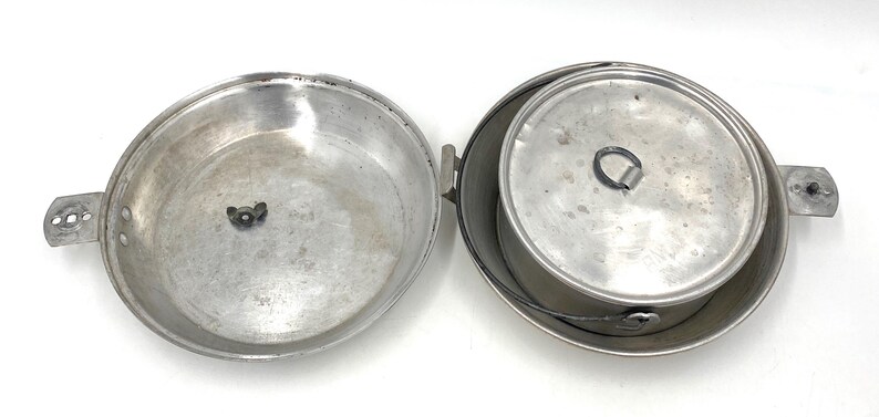 Op de afbeelding: Een open, vintage aluminium kampeerkookset. De set bevat een grote pan met een handvat en een kleinere pot met een deksel en een draadhandvat. Ideaal voor buiten koken en kamperen.