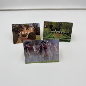 May include: Three postcards featuring historical sites. The first postcard shows a brick house with a green lawn and a walkway lined with flowers. The second postcard shows a formal garden with a red brick path and a white fence. The third postcard shows a group of men in colonial era clothing marching in a field.