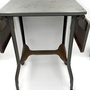May include: A gray metal drop-leaf table with a rectangular top and two hinged side panels. The table features four curved legs, each ending in a small caster wheel, and a horizontal support bar connecting the legs. The metal surface shows wear.