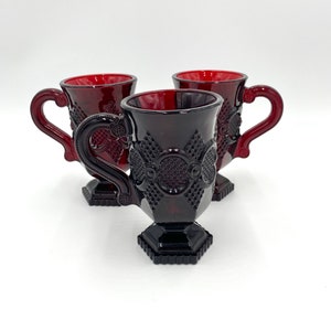 Puede incluir: Tres tazas de vidrio rojo con base y diseño decorativo. Las tazas tienen un asa y están hechas de vidrio rojo rubí.