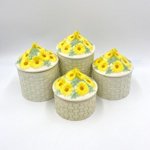 May include: Set of four ceramic canisters with yellow daisy flower designs and green wicker-like patterns. The canisters have lids with the same daisy flower design.